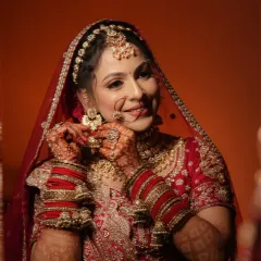 A candid moment of a bride getting ready. This image captures the rich detail of her red and gold embroidered lehenga, traditional chooda, and the warm tones of her makeup.