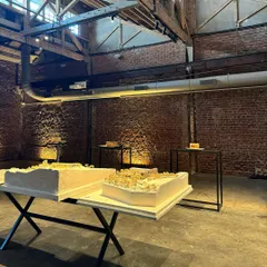 Architectural models on display within the main hall of the restored ice factory. The interplay of focused lighting on the exposed brick walls highlights the building's original texture, creating a dramatic gallery setting.