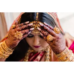 A close-up of the bride's final touches. Her hands, adorned with henna and gold rings, adjust the intricate temple jewelry, a key element of the South Indian bridal aesthetic.