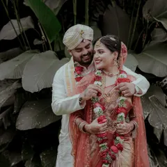 The groom helps the bride with her garland in this candid wedding portrait. The natural, leafy background contrasts beautifully with their traditional wedding attire.