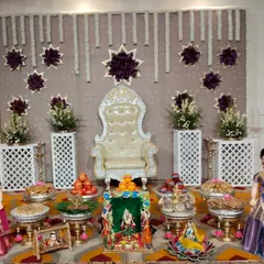 A complete and elegant baby shower setup with a beautiful white throne. The backdrop features hanging jasmine garlands and purple floral stars, complemented by traditional dolls and brass lamps.