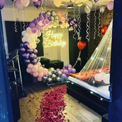 For a truly romantic birthday surprise, I transformed this room with a dreamy setup. It features a beautiful crescent moon balloon arch in shades of purple and pink, a 'Happy Birthday' neon sign, and a delicate canopy over the bed with fairy lights. The floor covered in rose petals completes the magical look.