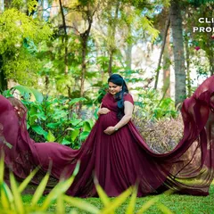 A serene portrait focusing on the mother-to-be cradling her bump. The elegant drape of the maroon gown adds a rich texture and color to the natural surroundings.