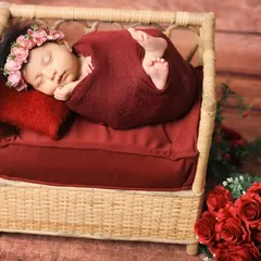 This is the 'tucked-in' pose, showcasing the baby's tiny feet. The newborn is swaddled in a deep red wrap on a miniature rattan bed, surrounded by roses, creating a rich and romantic feel.