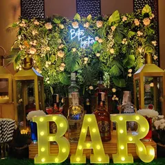 The perfect backdrop for a glamorous party. This bar setup features a lush green and white floral wall with a "Better Together" neon sign, complemented by large golden lanterns and a glowing 'BAR' marquee sign.