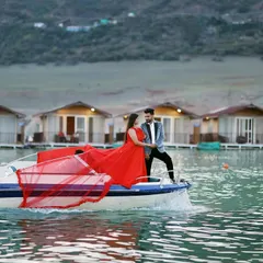 A dynamic shot of a speedboat creating a wake, with the couple enjoying the ride. The woman's red gown adds a splash of color and drama against the floating cottages of Tehri.