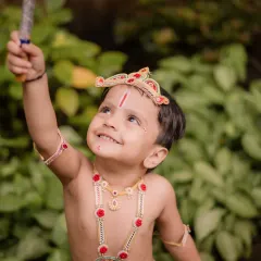 Full of joy and mischief, this little Krishna looks up with a bright smile while holding a prop, capturing the playful spirit of childhood.