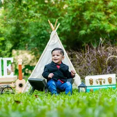 A one-year-old boy looking sharp in a little blazer, smiling for his pre-birthday photoshoot in a classic outdoor traveler theme setup.