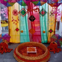 A vibrant Haldi ceremony space featuring a multi-colored backdrop with hanging kites and umbrellas. The 'urli' is filled with flower petals, ready for the rituals.