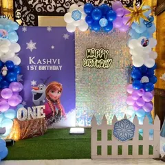 A magical first birthday with a Disney's Frozen theme, featuring a shimmer wall, character cut-outs of Anna and Elsa, and balloon pillars in icy blue, purple, and white.
