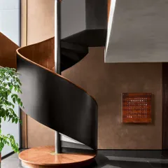 The spiral staircase at the Baid Residence is a study in elegant contrasts. The warmth of the terracotta-painted wall and the wooden steps is balanced by the cool, sleek finish of the blackened steel structure and honed black granite floor.