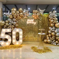 I handle decorations for major milestone celebrations, like this 50th birthday party. The setup features a glamorous gold shimmer wall for photos, large light-up marquee numbers, a custom 'Happy Birthday' neon sign, and a full balloon arch.