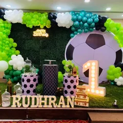 A football-themed first birthday party setup. This includes a green grass-like floor, a large football backdrop, themed pedestals, and a balloon arch in team colors.