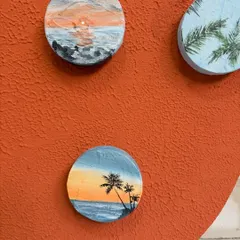 A close-up of some of the miniature travel paintings. Here you can see a sunset over the ocean and a beach with palm trees, each painted on a small wooden circle.