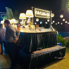 Our portable cocktail bar setup is perfect for outdoor events. This one features a "Delhi Cocktails Night" neon sign, elegant lamps, and paper lanterns hanging in the background.