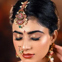 A detailed close-up of bridal makeup. This shot highlights the shimmering gold eyeshadow, the perfectly defined brows, and the intricate details of the maang tikka and nose ring.