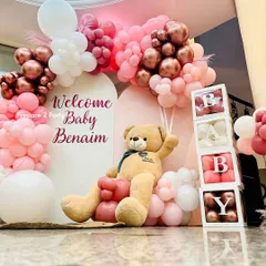 A beautiful and modern welcome home decoration for a baby girl. This setup uses a stylish arch backdrop, a lush balloon arrangement in shades of pink, rose gold, and white, along with "BABY" blocks and a large, cuddly teddy bear.