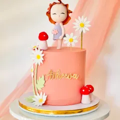 A sweet and simple cake with a girl figurine, mushrooms, and daisy decorations. The peach-colored buttercream and gold name topper make it a lovely birthday choice.