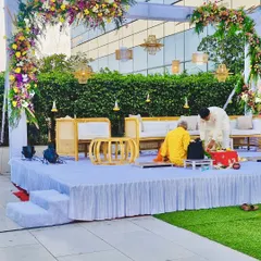 A modern and minimalist vidhi mandap for an outdoor daytime ceremony. The setup features a beautiful floral arch, comfortable cane furniture for the rituals, and elegant hanging lamps.