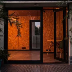 The same OSB-clad workspace viewed from the outside at dusk. The warm interior light creates an inviting glow, turning the small office into a welcoming beacon.