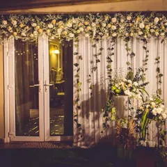 An elegant floral backdrop designed for an evening event. A cascade of white flowers and trailing ivy over sheer curtains creates a soft, romantic photo wall, complemented by modern floral stands.