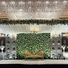 An absolutely stunning stage setup at Crowne Plaza, Okhla. A massive crystal chandelier hangs above, complementing the white floral wall, intricate side panels, and elegant white drapes.