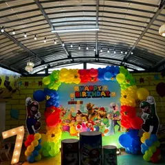 This Super Mario theme brings the iconic video game to life with a full backdrop of the Mushroom Kingdom. The setup is framed by a rainbow balloon arch and features character cutouts of Mario and Luigi, along with a light-up marquee number '7'.