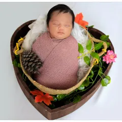 A beautifully composed shot of a newborn swaddled in a dusty rose wrap, nestled inside a heart-shaped wooden basket. The floral and pinecone accents add a natural, rustic touch.