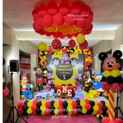 This is a complete Mickey Mouse themed first birthday party I created. The setup includes a personalized backdrop with the child's photo, multiple character cutouts, and coordinated red, yellow, and black balloon decorations for a fun and immersive experience.