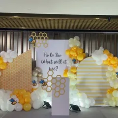 A unique and adorable 'He or She, What will it bee?' gender reveal theme. The backdrop features a honeycomb pattern with yellow, white, and gold balloons and cute bee accents for a playful touch.