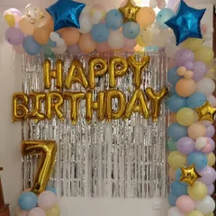 A classic "Happy Birthday" setup for a 7th birthday at home. It features a silver tinsel curtain, gold foil letters, and a frame of pastel-colored balloons.