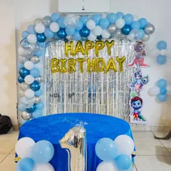 A first birthday party setup with a cartoon character theme. The decor includes a blue and silver balloon arch, a silver tinsel backdrop, and a decorated table for the cake.