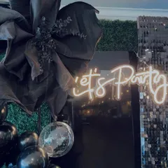 A close-up shot of the "Let's Party" neon sign, showing how it reflects beautifully against the dark, glamorous decor elements like the sequin wall and large black bow.