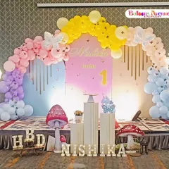 We love creating magical first birthday parties like this butterfly garden theme. The setup includes a full stage with a pastel rainbow balloon arch, custom name backdrop, illuminated number one, and whimsical props like giant mushrooms and flower lights.