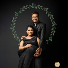 A formal maternity portrait against a solid black background. The couple is dressed in elegant black attire, and a simple digital wreath was added to frame them, creating a classic and sophisticated look.