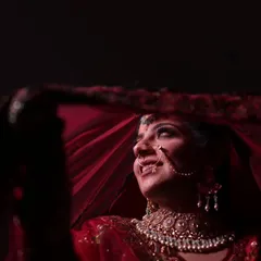 A classic bridal shot, capturing the bride's joyful smile as she peeks out from under her red veil. The lighting highlights her features and the sparkle of her diamond necklace, creating a portrait that is both traditional and full of life.