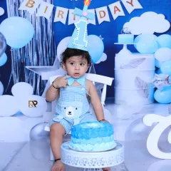 A sweet moment during a cake smash session, where the birthday boy sits calmly in front of his cake. The blue and white setup with a "Happy Birthday" banner creates a classic party atmosphere.