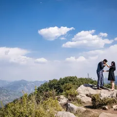 A romantic gesture on a mountaintop, with him kissing her hand against a vast blue sky filled with fluffy clouds. This wide shot captures a feeling of being on top of the world.