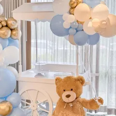 We use charming props to enhance our themes. Here, a large, cuddly teddy bear sits next to a white cart, which is perfect for holding gifts or a cake, all tied together with a blue, white, and gold balloon arrangement.