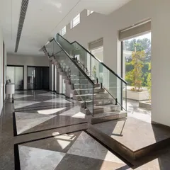 Sunlight streams into this hallway, creating a beautiful play of shadows across the geometric inlaid marble floor. The modern glass-railed staircase appears to float, enhancing the sense of light and space.