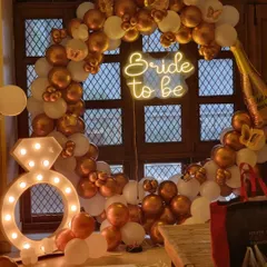 Celebrate the bride-to-be in style. This chic bachelorette party decoration features a "Bride to be" neon sign on a gold and white balloon arch, complete with a large light-up diamond ring prop for fun photos.