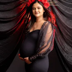 A powerful solo portrait with a touch of red. The mom-to-be wears a striking red floral crown that contrasts beautifully with her black dress and the dark, draped background.