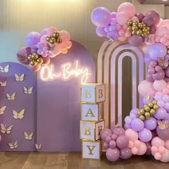 A stunning "Oh Baby" shower backdrop in shades of lavender and pink. The decor includes butterfly accents, gold details, "BABY" blocks, and a neon sign for a touch of modern glam.