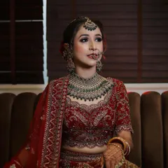 A full-length view of the bride seated, showcasing her complete look. The makeup is perfectly balanced with her elaborate red lehenga and heavy jewelry, creating a regal and timeless bridal appearance.