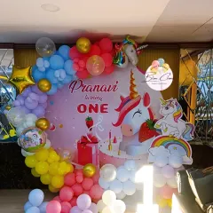 A magical unicorn-themed first birthday party. The decor features a custom backdrop with unicorns and rainbows, surrounded by a colorful pastel balloon garland.