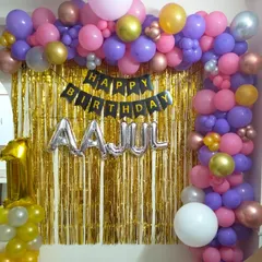 A first birthday party decoration designed for a home setting. It features a gold fringe backdrop, a "Happy Birthday" banner, and a colorful balloon arch.