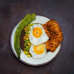 A healthy and high-protein meal with grilled chicken breast, sunny-side-up eggs, quinoa, and green beans. The dark, dramatic lighting and top-down view create a powerful and appetizing fitness meal shot.