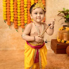 A classic portrait of a baby dressed as little Krishna for Janmashtami, holding his flute. The setup is decorated with marigold flowers for a festive feel.