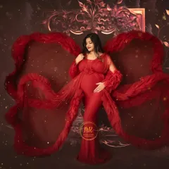 A dramatic solo portrait in a deep red gown with feathered details. The fabric is styled to create a wing-like effect against a rich, textured backdrop, perfect for a bold pregnancy announcement.