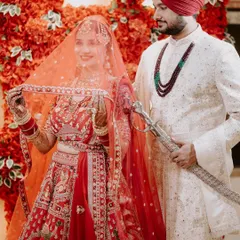 A classic portrait of a Sikh couple on their wedding day. The groom, holding the kirpan, stands proudly with his bride, who lifts her veil with a smile, against a stunning red floral backdrop.